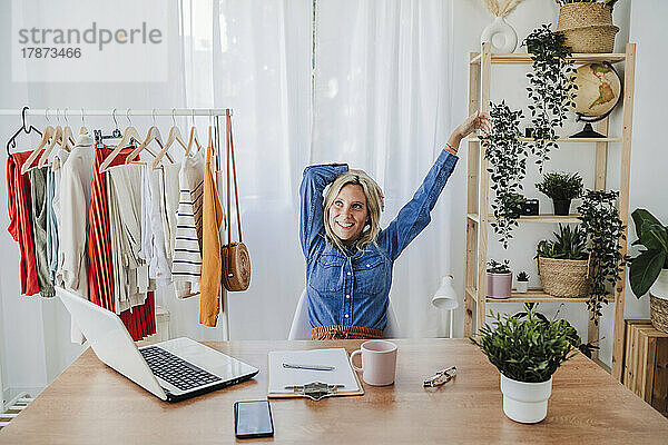 Lächelnde Geschäftsfrau mit erhobenen Armen sitzt am Schreibtisch im Heimbüro