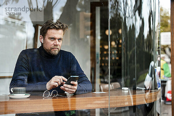 Reifer Geschäftsmann mit Mobiltelefon sitzt im Café und sieht durch die Glasscheibe