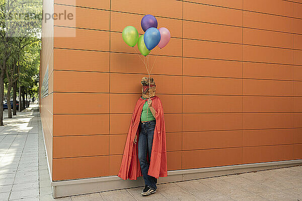 Junge Frau mit Krokodilmaske und Umhang steht mit bunten Luftballons vor der Wand