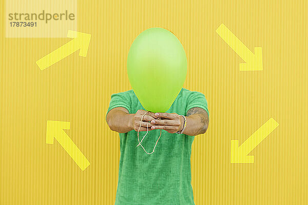 Young man covering face with green balloon in front of arrow symbols on yellow wall