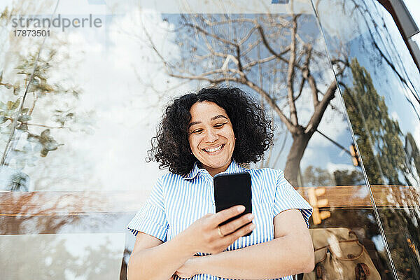 Glückliche Frau  die ihr Mobiltelefon benutzt und vor Glas steht