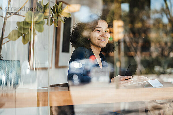 Lächelnde Frau mit Smartphone  gesehen durch das Glas eines Cafés