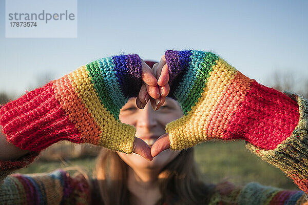 Lächelnde Frau mit Regenbogenhandschuhen gestikuliert an einem sonnigen Tag in Herzform