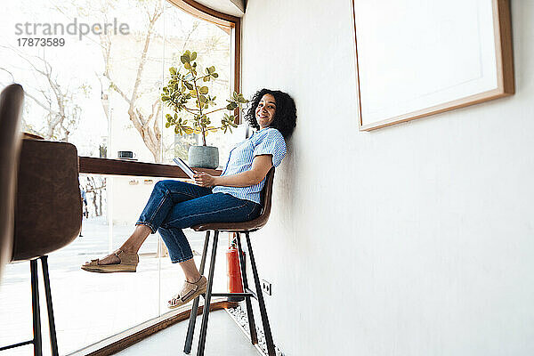 Glückliche Frau mit Tablet-PC sitzt auf einem Stuhl am Fenster im Café