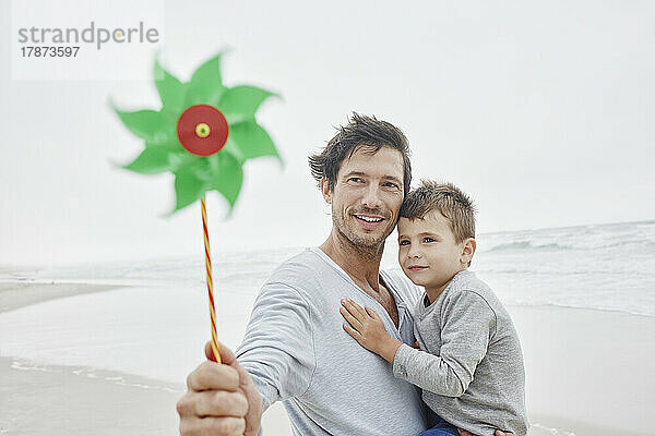 Vater trägt Sohn am Strand und hält grünes Windrad in der Hand