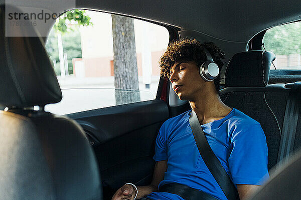 Tired young man wearing wireless headphones sleeping in car on road trip