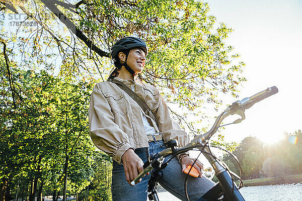 Lächelnde Frau mit Smartphone sitzt auf Fahrrad im Park