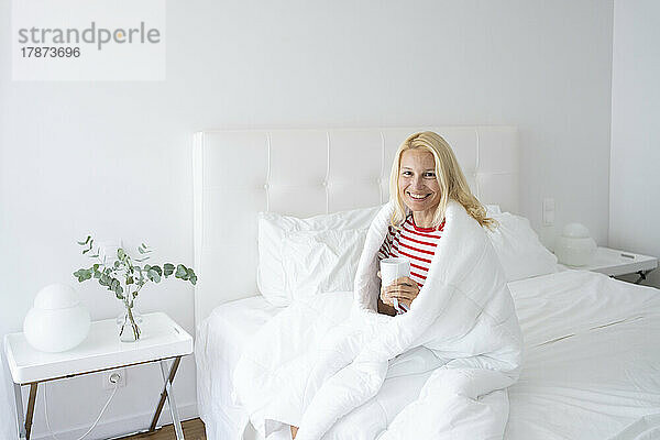 Lächelnde Frau  in eine Decke gehüllt  trinkt Tee im Bett