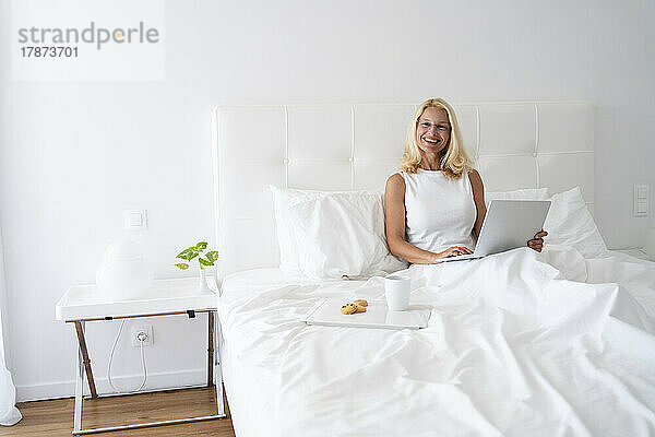 Glückliche Frau mit Laptop sitzt neben dem Frühstückstablett auf dem Bett