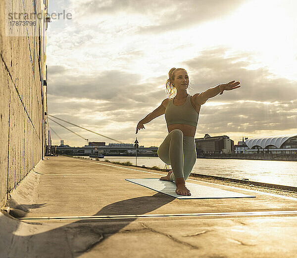 Lächelnde Frau mit ausgestreckten Armen praktiziert Yoga auf der Promenade bei Sonnenuntergang