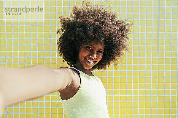 Lächelndes Mädchen mit Afro-Frisur steht vor gelber Wand