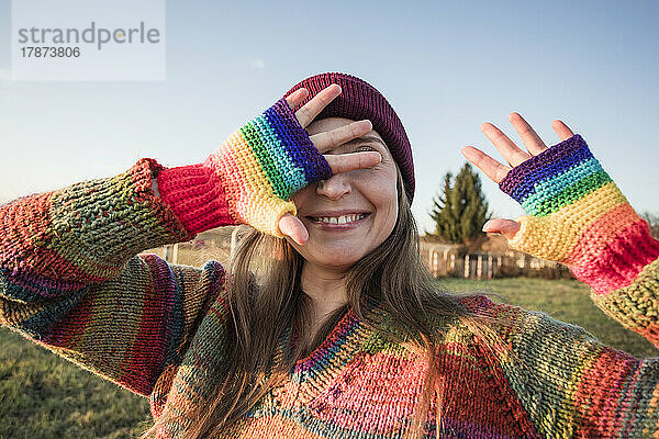 Glückliche Frau trägt Regenbogenhandschuhe  die ihr Gesicht mit den Händen bedecken