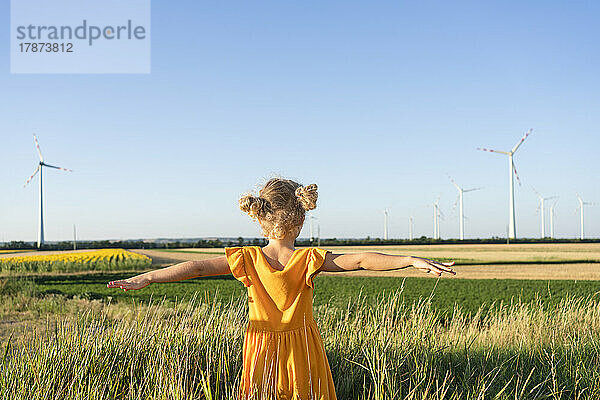 Mädchen mit ausgestreckten Armen steht auf dem Feld und blickt auf Windkraftanlagen