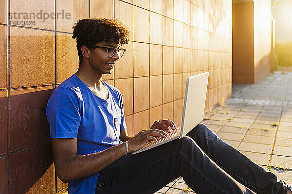 Lächelnder Mann  der Laptop zum Lernen benutzt und an der Wand sitzt