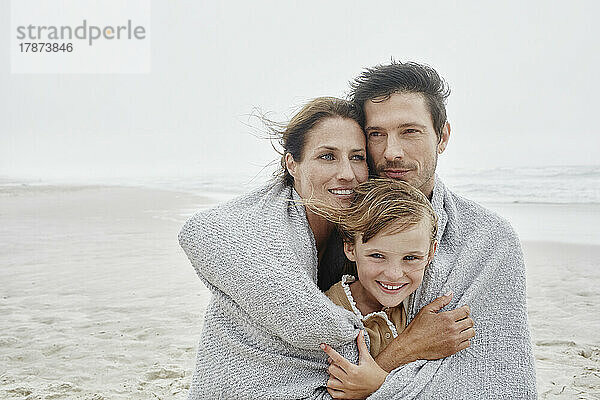 Mann und Frau stehen mit Tochter am windigen Strand  in eine Decke gehüllt
