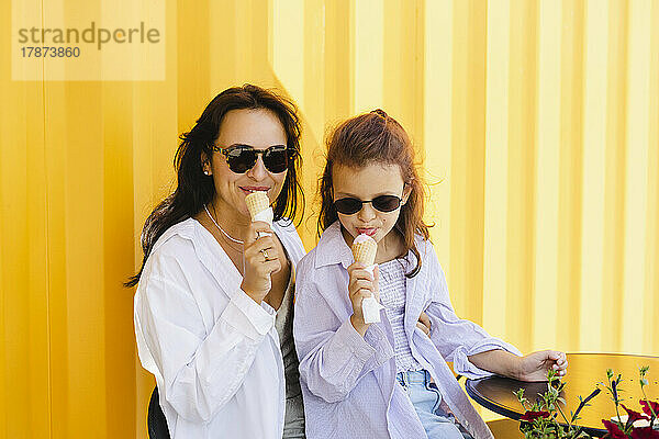Mutter und Tochter mit Sonnenbrille essen Eis vor gelber Wand