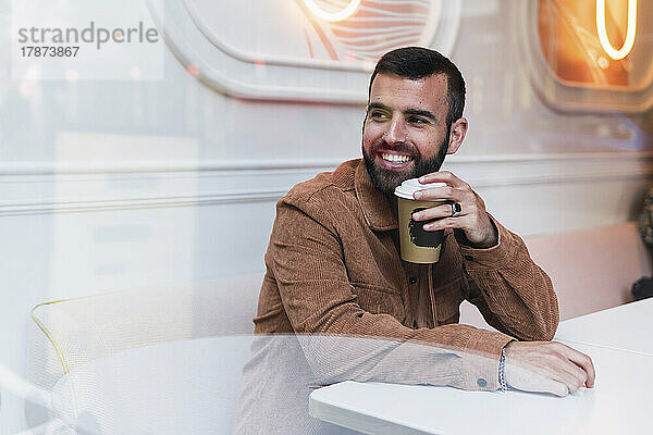 Lächelnder junger Mann mit Einwegkaffeetasse sitzt am Tisch im Café