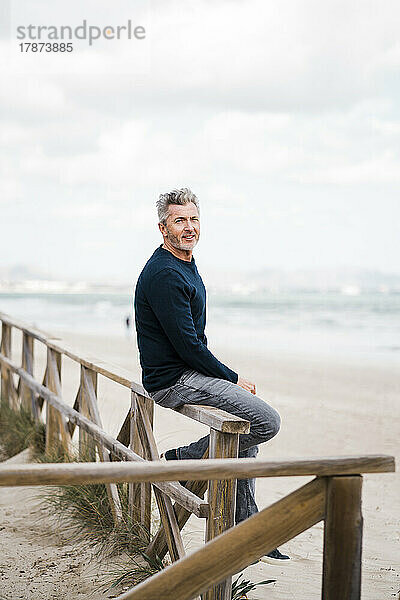 Glücklicher reifer Mann sitzt auf dem Geländer am Strand