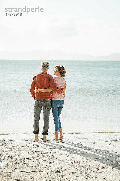 Älteres Paar mit umeinander geschlungenen Armen steht am Ufer des Strandes