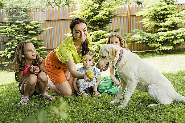 Lächelnde Frau zeigt Hund von Töchtern im Hinterhof den Ball