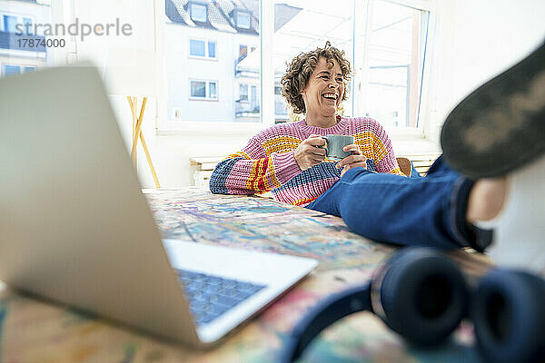 Happy woman holding coffee cup sitting with feet up on table at home
