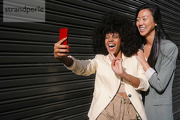 Fröhliche Frau zeigt ein Friedenszeichen und macht an einem sonnigen Tag ein Selfie mit einer Freundin in der Nähe des Fensterladens