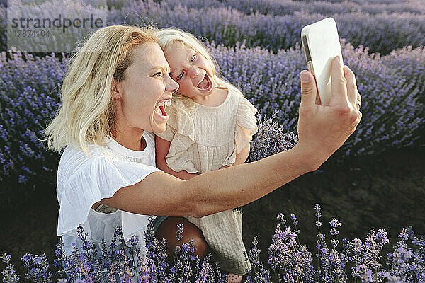 Fröhliche Mutter macht mit ihrer Tochter ein Selfie mit dem Handy inmitten von Lavendelblüten