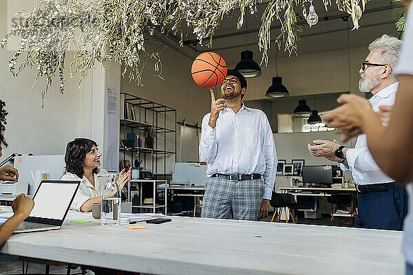 Verspielter Geschäftsmann dreht Basketball am Finger  während Kollegen im Büro jubeln