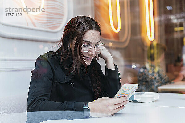 Smiling young woman using smart phone sitting at cafe