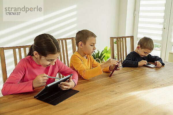 Happy girl using tablet PC sitting by brothers with technologies at home