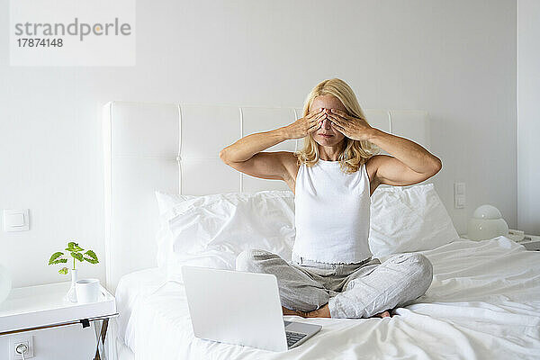 Gestresste Frau verdeckt die Augen und sitzt am Laptop im Bett