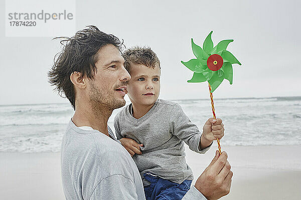 Vater trägt Sohn am Strand und hält grünes Windrad in der Hand