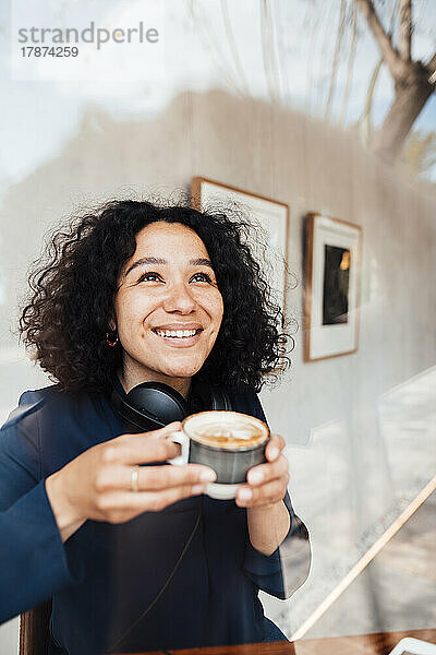 Glückliche Frau mit lockigem Haar und Kaffeetasse  gesehen durch Glas im Café