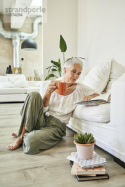 Frau hält einen Becher und liest ein Buch auf dem Sofa