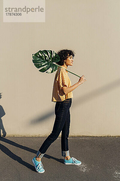 Frau hält Monstera-Blatt und steht an einem sonnigen Tag vor der Wand