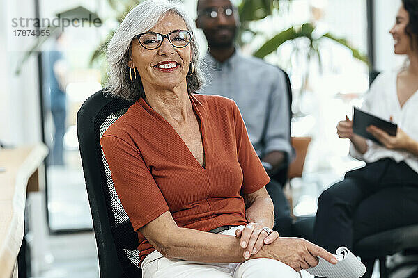 Lächelnde ältere Geschäftsfrau sitzt auf einem Stuhl am Arbeitsplatz