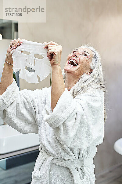 Reife Frau mit Gesichtsmaske lacht zu Hause im Badezimmer