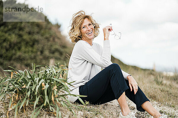 Glückliche reife Frau mit Brille sitzt neben einer Pflanze am Strand