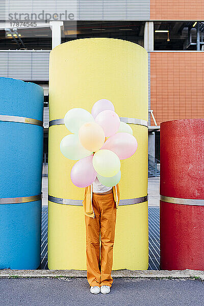 Junge Frau bedeckt Gesicht mit bunten Luftballons vor Rohren