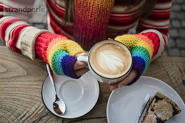 Hände einer Frau mit Regenbogenhandschuhen  die eine Kaffeetasse am Tisch hält
