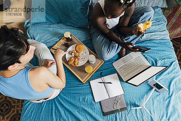 Woman holding mobile phone having breakfast with girlfriend on bed