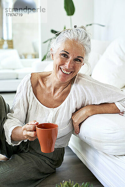 Lächelnde Frau hält eine Kaffeetasse in der Hand und lehnt auf der Couch