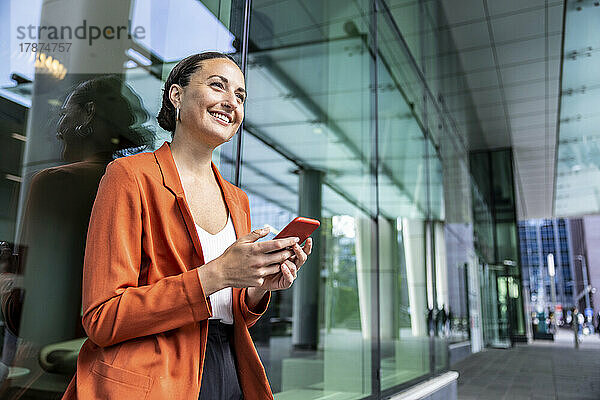 Glückliche Geschäftsfrau hält Mobiltelefon an Glaswand gelehnt