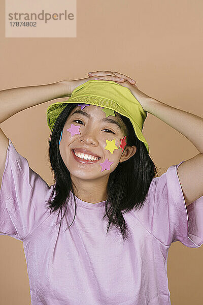 Smiling woman with multi colored stars on face wearing bucket hat against brown background