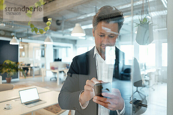 Reifer Geschäftsmann surft im Internet über sein Mobiltelefon und sieht durch Glas im Büro