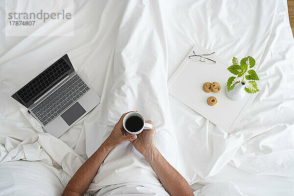 Hände eines Mannes  der mit schwarzem Kaffee und Laptop auf dem Bett sitzt