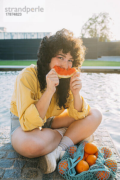 Frau mit Orangen im Netzbeutel isst Wassermelone am Teich