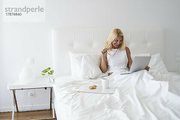 Lächelnde Frau  die beim Frühstück im Bett am Laptop arbeitet
