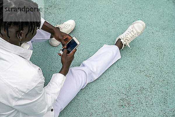 Junger Mann schreibt Textnachrichten per Smartphone und sitzt auf dem Fußweg
