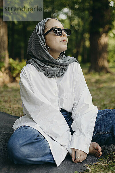 Junge Frau mit Sonnenbrille und Hijab sitzt im Schneidersitz im Park